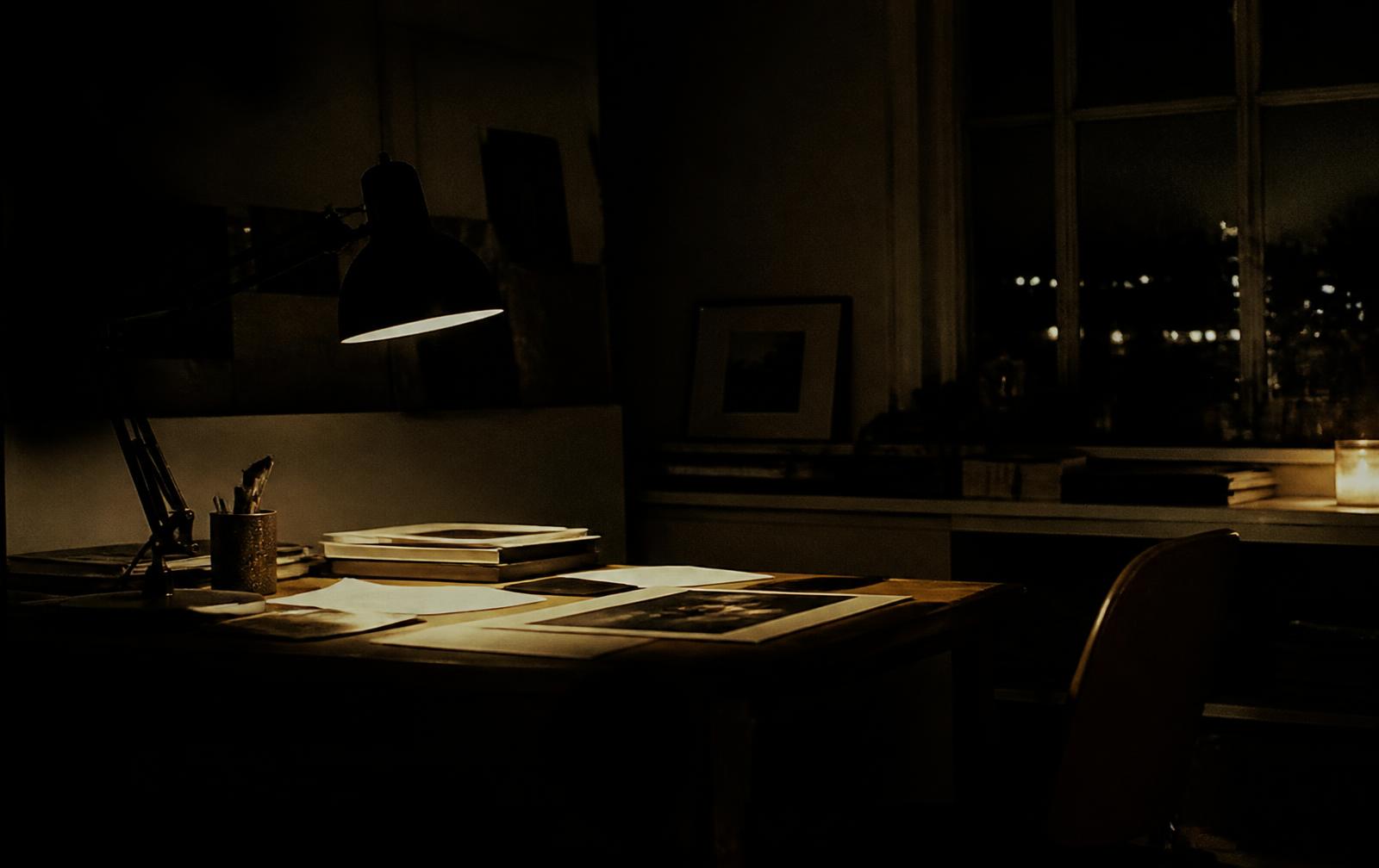 A dim desk at night with a lamp, papers and prints