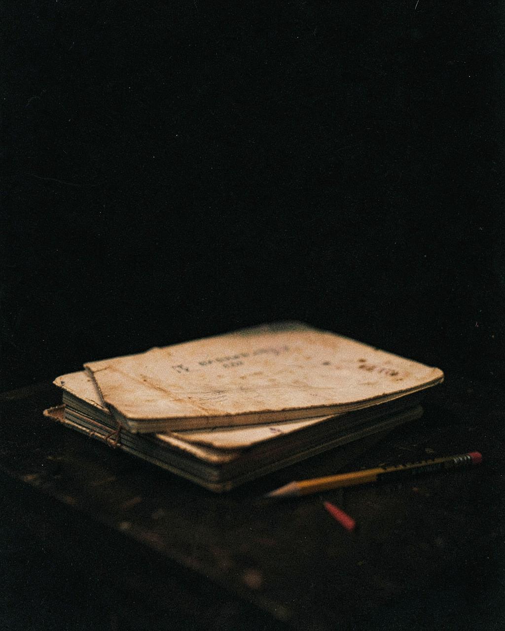 A stack of worn notebooks and a pencil on a dark table